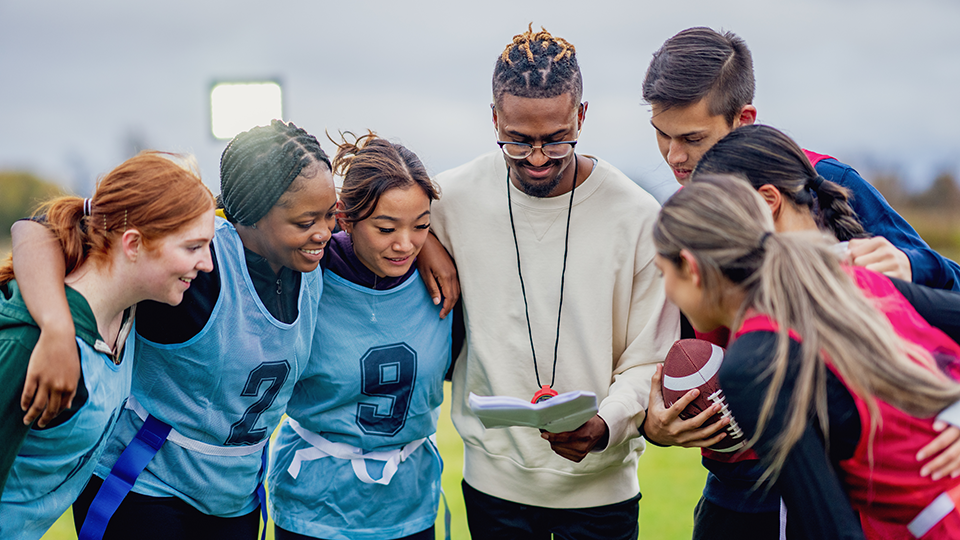 A coach holding a clipboard reviews plays with a diverse group of young athletes wearing flag football pinnies on an outdoor field