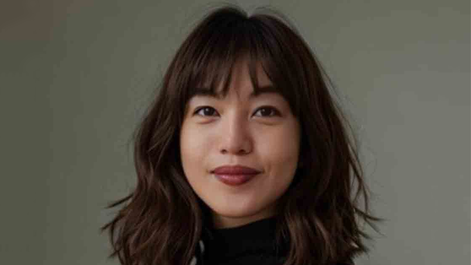 Portrait of a woman with shoulder-length wavy brown hair and bangs, wearing a black top, smiling softly against a neutral gray background